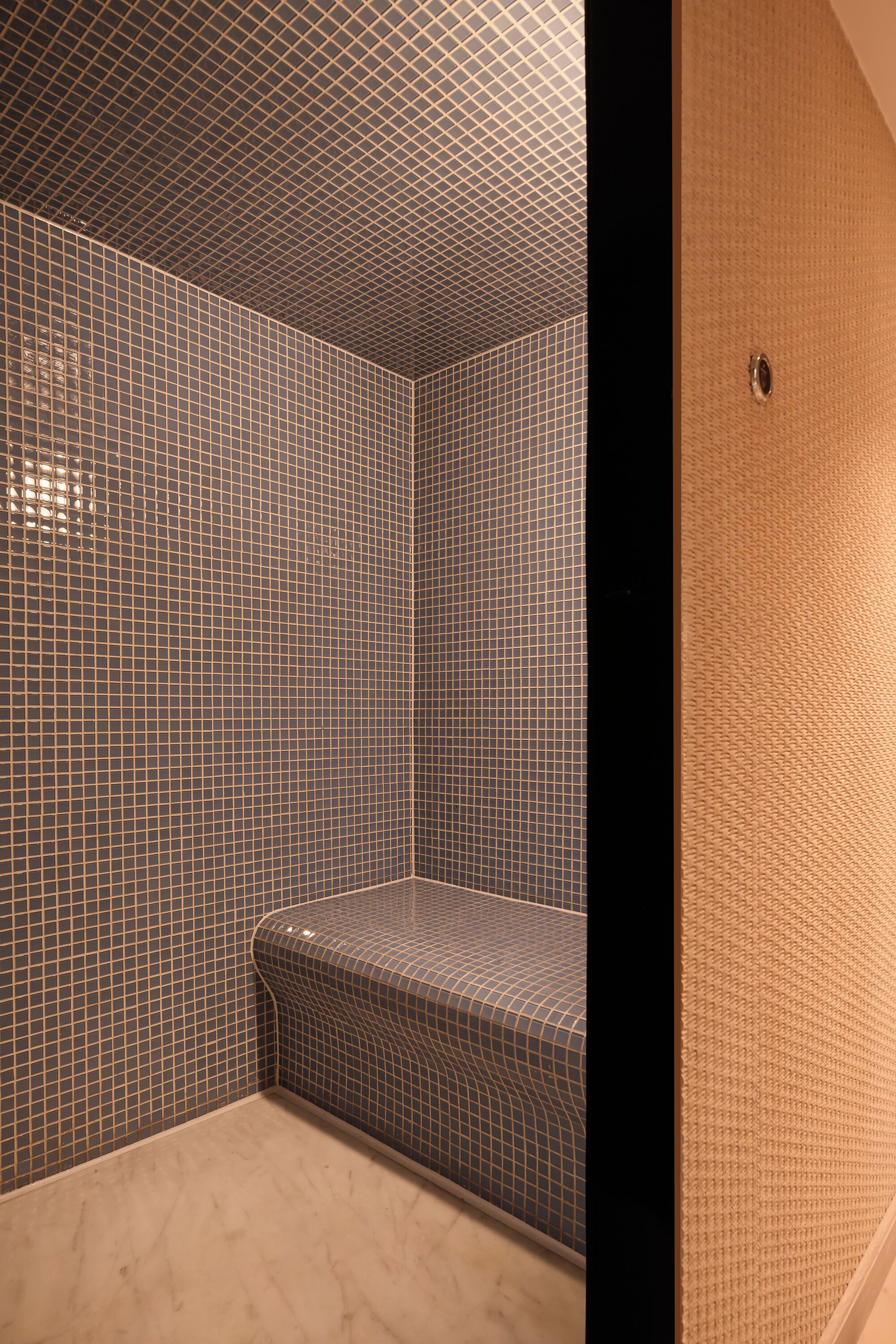 A bathroom with a tiled floor and walls, a bench, and a door with a textured surface.