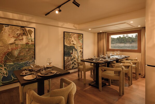 A dining area with a table set for six, surrounded by chairs, with large paintings on the walls and a window with a view of the water. - Le Fayan Suites Nile Cruise - Mondays from Luxor & Fridays from Aswan. (Luxor)