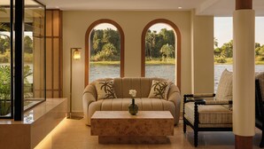 A living room with a sofa, a coffee table, and a chair. There are two arched windows with a view of greenery and water. - Le Fayan Suites Nile Cruise - Mondays from Luxor & Fridays from Aswan. (Luxor)