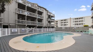 Une piscine dotée d’une terrasse en bois, entourée d’une clôture blanche et bordée d’un bâtiment comportant plusieurs balcons.