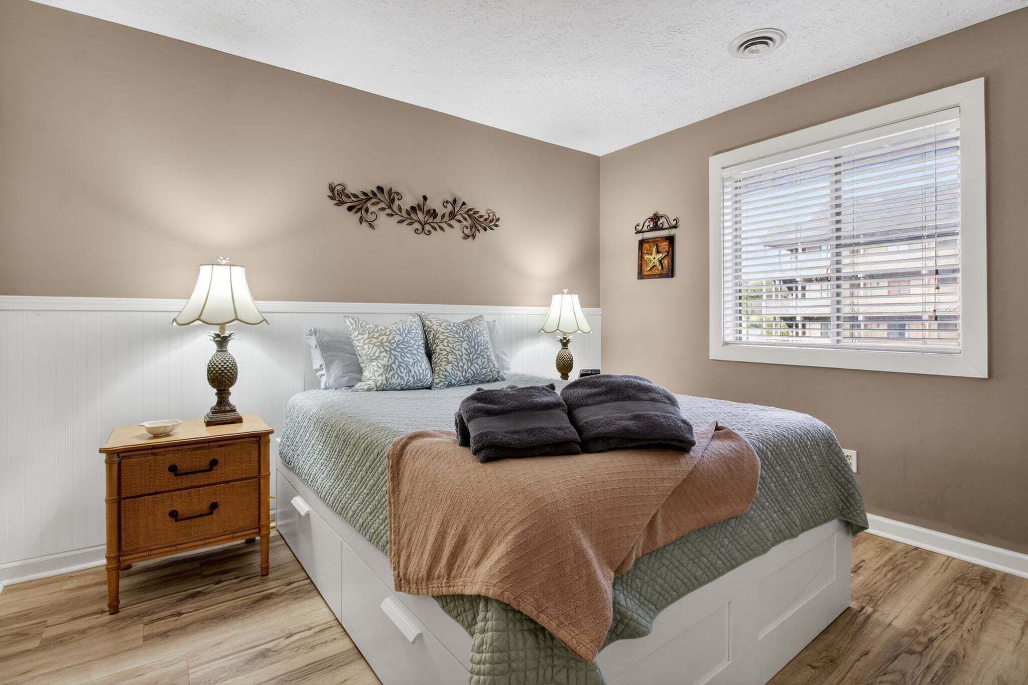 A bedroom with a bed, two bedside tables, a window with blinds, and a decorative wall piece.