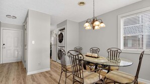 Un coin repas avec une table en verre et des chaises en métal, une cuisine en arrière-plan et une fenêtre avec des stores.