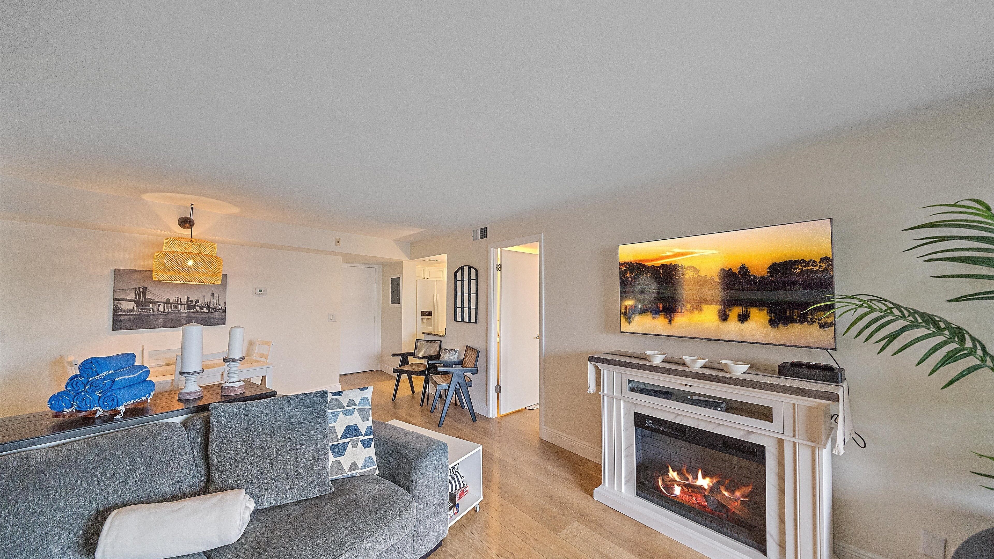 A modern living room with a fireplace, a dining area, and a kitchen visible in the background.