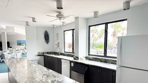 A modern kitchen with a marble countertop, dark cabinets, a built-in dishwasher, and a large window offering a view of palm trees. - Sole Retreat 2 Bedroom Condo (Siesta Key)