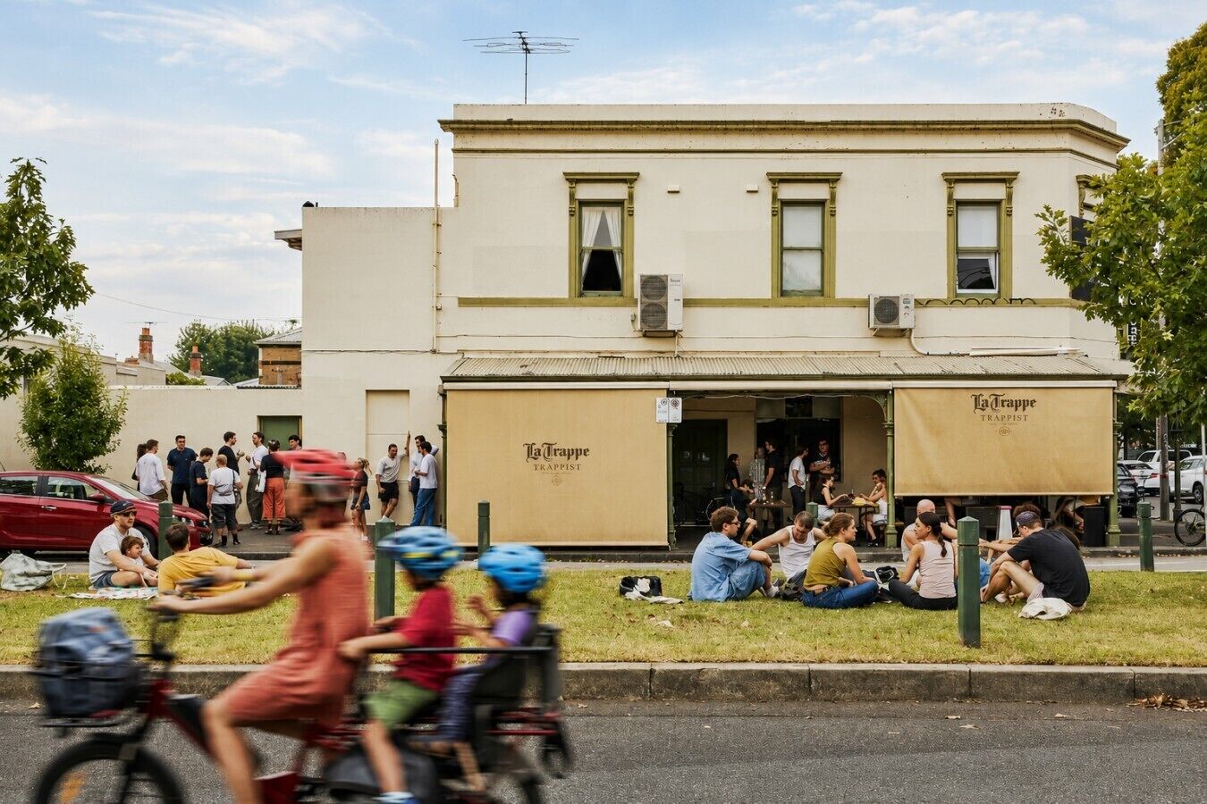 A building with a sign reading 'La Trappe Trappist' and a group of people sitting on the grass in front.
