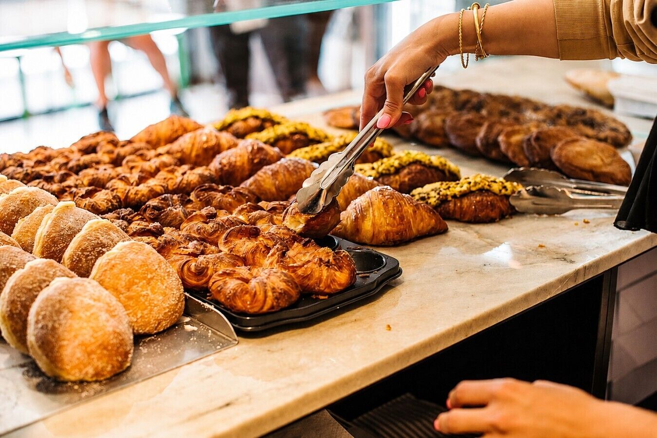 Una varietà di prodotti da forno esposti, tra cui croissant e altri dolci, con una persona che usa delle pinze per sceglierne uno.