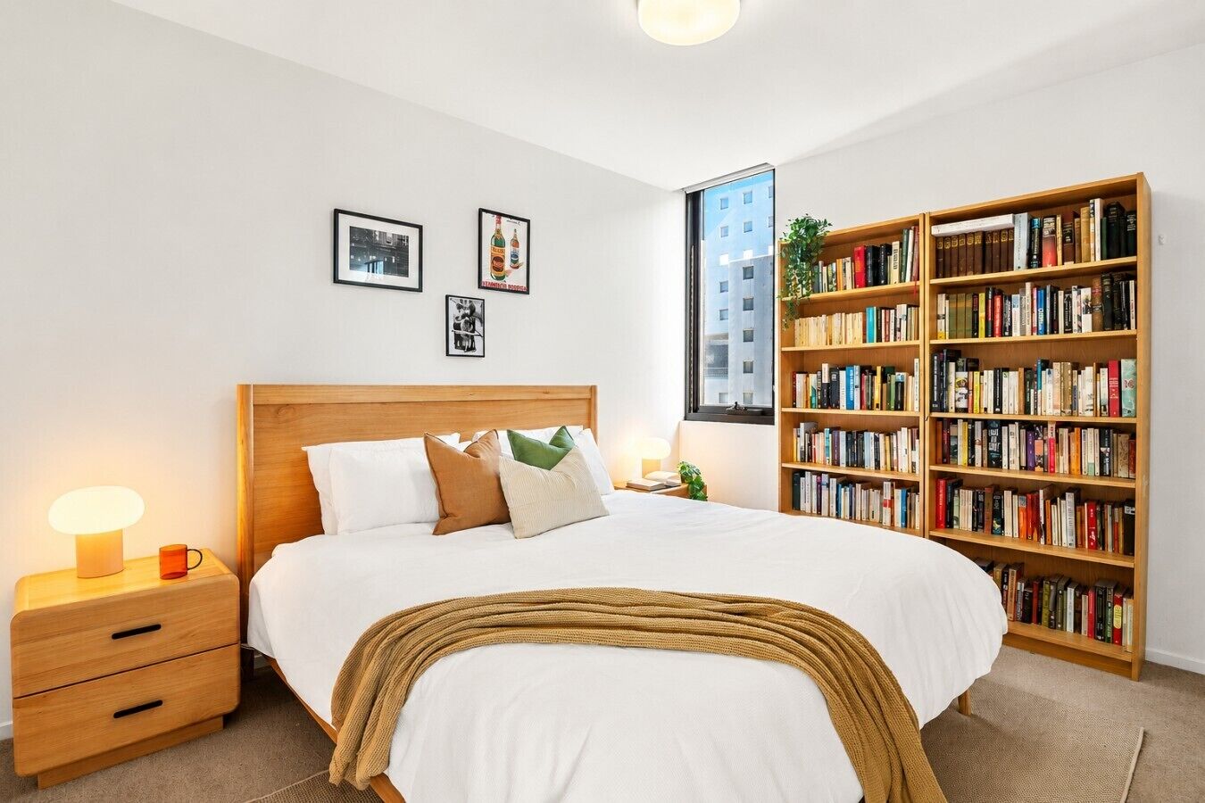 Een slaapkamer met een groot bed, een nachtkastje met een lamp, een boekenkast en ingelijste foto's aan de muur.