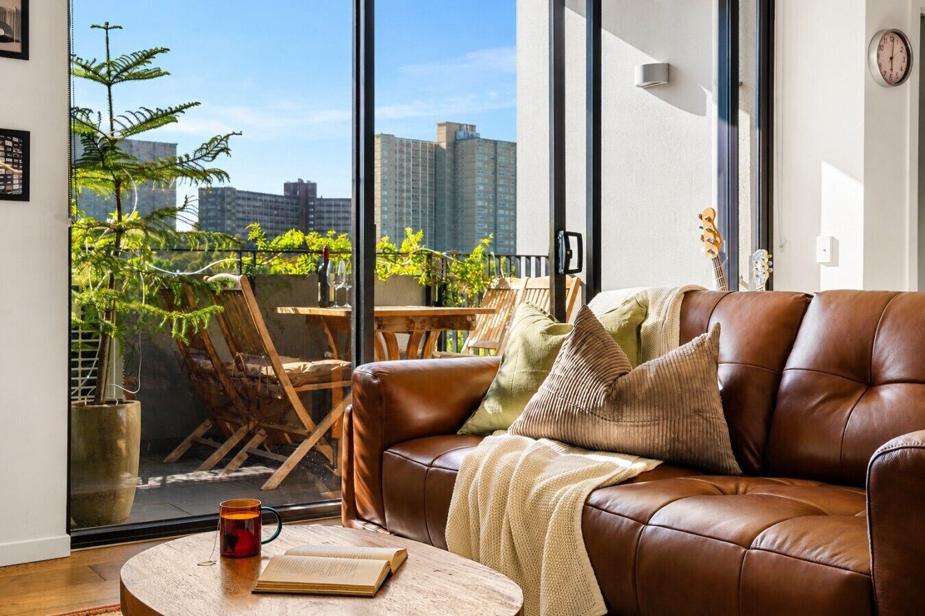 A cozy living room with a leather sofa, a coffee table with a book and a mug, and a view of a balcony with plants and a cityscape.