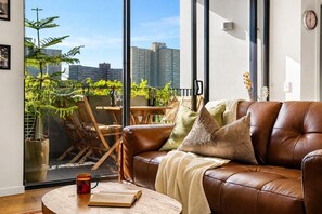 Ein gemütliches Wohnzimmer mit einer Ledersofa, einem Couchtisch mit Buch und Tasse, und Blick auf einen Balkon mit Pflanzen und Stadtansicht.