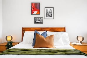 A neatly made bed with a wooden headboard, white bedding, and a green throw blanket. There are two bedside tables with lamps and a small plant on one. - The Italian Library: Amazing Views + Free Parking! (Carlton)