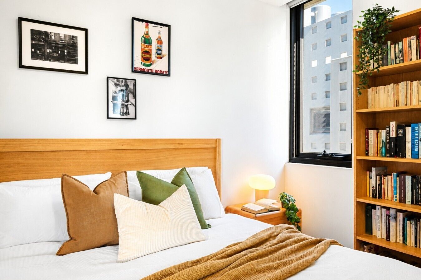 A bedroom with a wooden headboard, a bed with multiple pillows, a bedside table with a lamp, and a bookshelf filled with books.