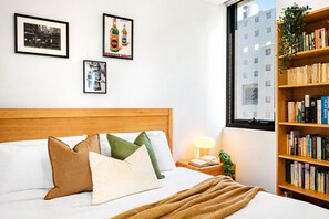 Ein Schlafzimmer mit Holzkopfteil, einem Bett mit vielen Kissen, einem Nachttisch mit Lampe und einem Bücherregal voller Bücher.