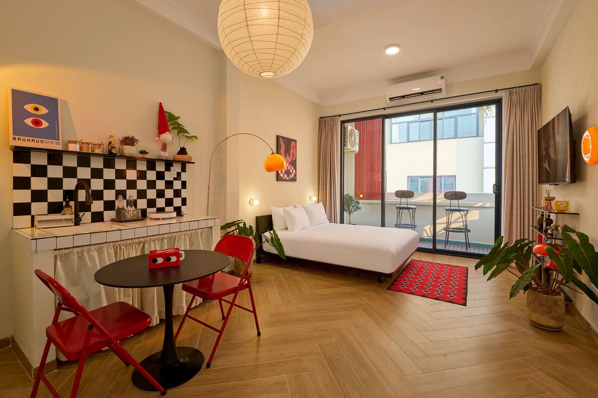 Moderni hotellihuone, jossa on ruutukuviollinen keittiön välitila, punaisilla tuoleilla varustettu ruokailutila, suuri sänky ja parveke, jossa on ulkokalusteita.