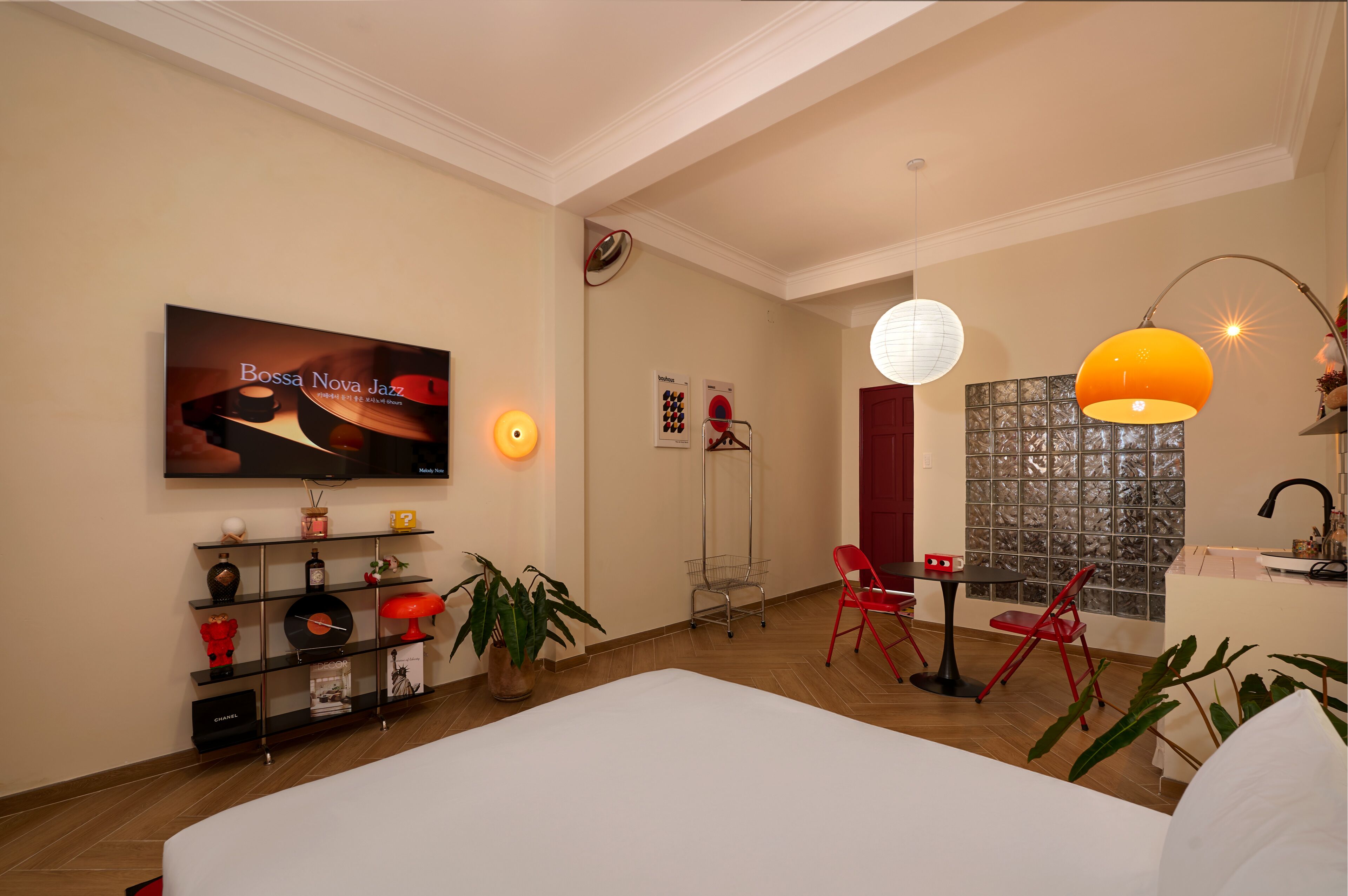A modern hotel room with a large bed, a TV displaying 'Bossa Nova Jazz', a red chair, a small table, and a decorative wall with a patterned window.