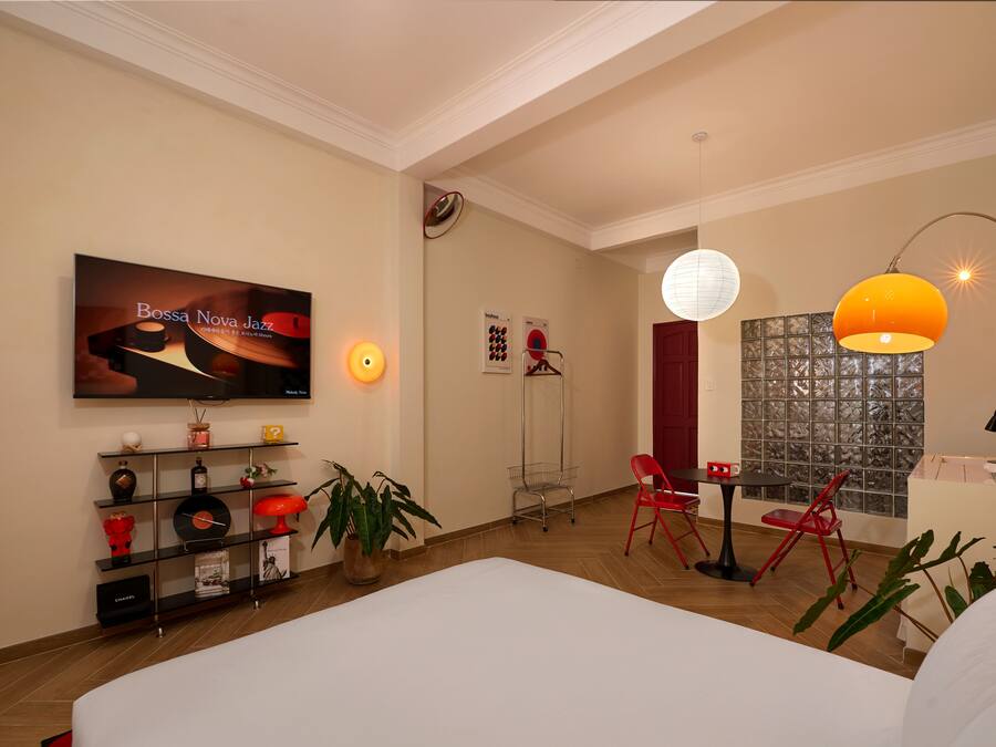 A modern hotel room with a large bed, a TV displaying 'Bossa Nova Jazz', a red chair, a small table, and a decorative wall with a patterned window.