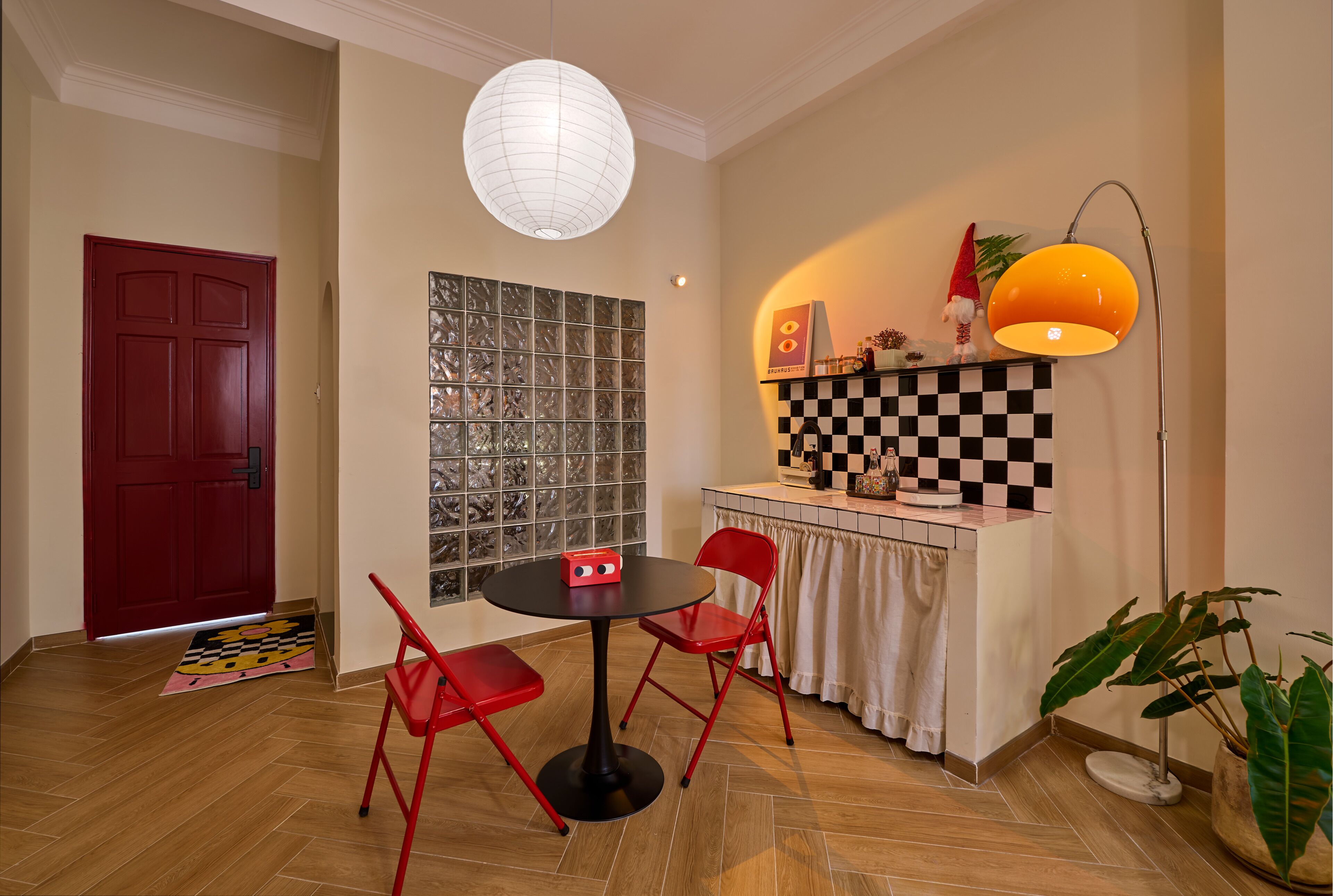 Una habitación con una puerta roja, una mesa redonda con dos sillas rojas, un mostrador a cuadros, una lámpara colgante blanca y una lámpara de pie.