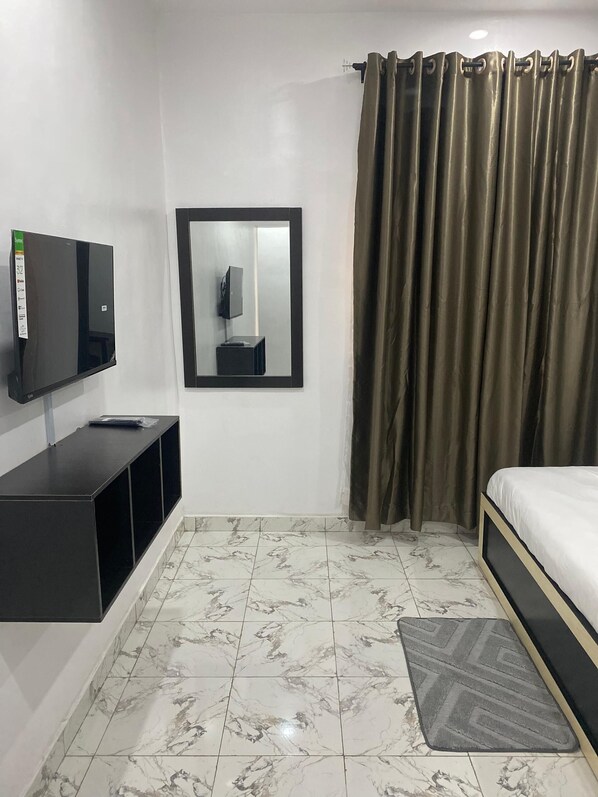 A hotel room with a bed, a TV mounted on the wall, a mirror, and a curtain. - ADONAI GUESTHOUSE (Agbara)