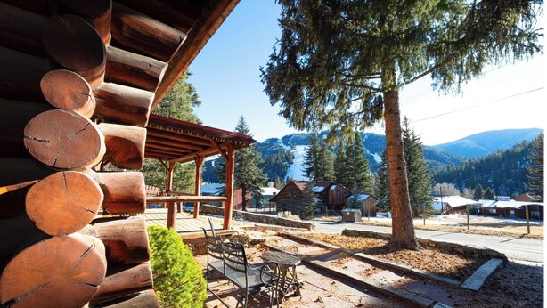 A wooden cabin with a covered patio, a metal table and bench, and a view of surrounding buildings and mountains. - A Cabin on High - In Town - Fireplace - Mountain Views (Red River)