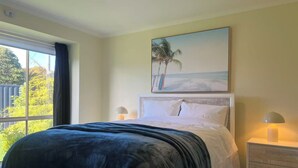 Una camera da letto con un letto, un comodino, un quadro raffigurante una spiaggia e una finestra vista verde.