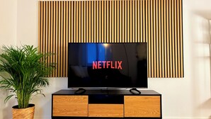 A modern living room with a flat-screen TV mounted on a wooden panel wall, a wooden cabinet with drawers, and a potted plant. - Vienna Homey Place - Spacious 70m2 Apartment (vie)