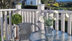 Una mesa de vidrio con una silla de mimbre, una planta pequeña en maceta y un florero con flores en un balcón con vistas a un cuerpo de agua y un edificio blanco.