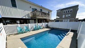 Ein rechteckiger Swimmingpool mit Holzterrasse, umgeben von einem weißen Zaun und blauen Adirondack-Stühlen.