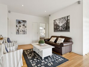 Ein modernes Wohnzimmer mit einem weißen Sofa, einem braunen Ledersessel und einem weißen Couchtisch. An den Wänden hängen zwei Bilder, eines zeigt Palmen und das andere eine Straßenszene.