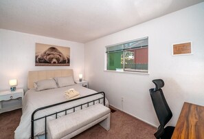 Ein Schlafzimmer mit einem Bett, Nachttischen, einem Fenster mit Jalousien, einem Sessel und einem Bärenbild an der Wand.