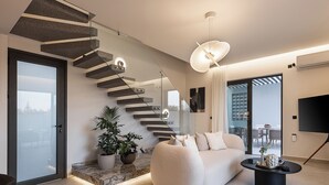 Un espacio habitable moderno con una escalera original, un sofá cómodo, una mesa de centro y un área de comedor visible a través de una puerta de cristal.