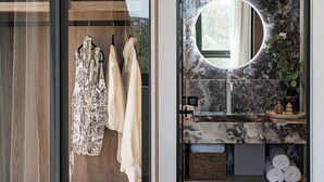 Un baño moderno con lavamanos de mármol, un espejo redondo y un vestidor tipo armario empotrado con ropa colgada.