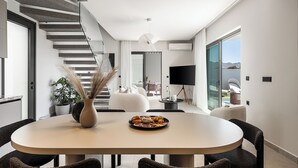 Un comedor moderno con una mesa redonda, sillas de ratán oscuro, un jarrón con plantas secas y un plato de pasteles. Una escalera con barandilla de cristal conduce a un balcón con vistas al exterior.