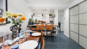 Ein moderner Essbereich mit einem langen Tisch, Stühlen und einem Blumenstrauß. Im Hintergrund ist eine Küchenzeile mit verschiedenen Gegenständen und einem Kühlschrank zu sehen.