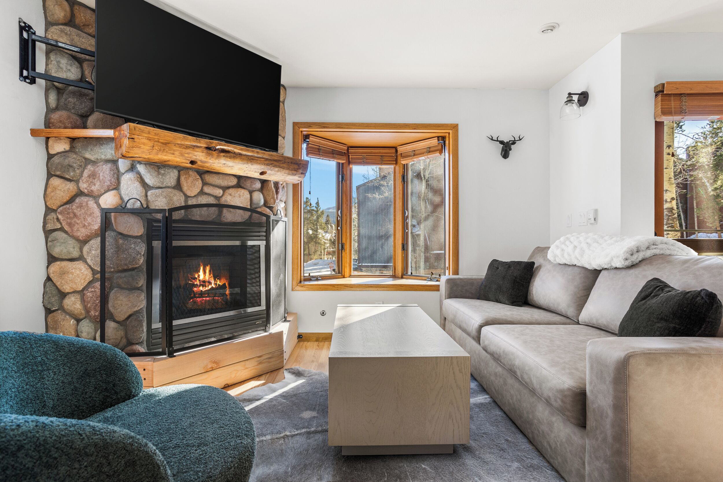 A living room with a stone fireplace, a flat-screen TV mounted above it, a large window with a wooden frame, and a stone wall.