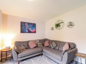 A modern living room with a grey sectional sofa, a round coffee table, a floor lamp, and a wall-mounted painting of a deer. - 6 Carn Dearg (Aviemore)
