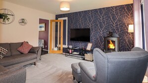 A living room with a sofa, armchair, flat-screen TV, fireplace, and a wall with a tree branch pattern. - 6 Carn Dearg (Aviemore)