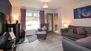 A modern living room with a sofa, armchair, and a fireplace. There is a window with curtains, a door leading to another room, and a painting on the wall. - 6 Carn Dearg (Aviemore)