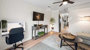 A modern living room with a black office chair, a wooden console table with plants, a black coffee table, and a ceiling fan. - Private Parking, Fire Ring, King Beds - Near Ark! (Dry Ridge)
