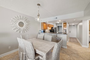 A dining area with wicker chairs, a wooden table, a sunburst mirror, and a kitchen in the background. - Boardwalk 585 by Liquid Life (Gulf Shores)