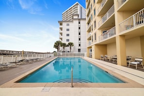 A swimming pool with lounge chairs and a wooden deck, surrounded by a multi-story building with balconies. - Boardwalk 886 by Liquid Life (Gulf Shores)