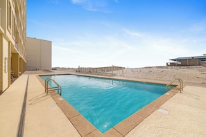A swimming pool with a clear blue water, surrounded by a sandy area and a building with a flat roof. - Boardwalk 886 by Liquid Life (Gulf Shores)