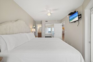 A hotel room with a large bed, a ceiling fan, a television mounted on the wall, and a view of the beach through the window. - Boardwalk 886 by Liquid Life (Gulf Shores)