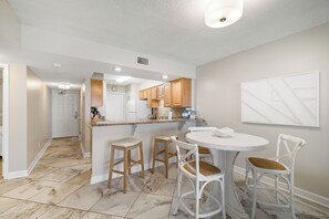 A modern kitchen with white cabinets, a central island, and a dining area with white chairs and a round table. - Boardwalk 886 by Liquid Life (Gulf Shores)
