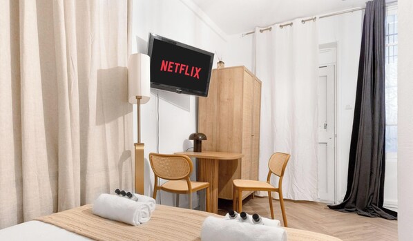 A hotel room with a bed, a TV mounted on the wall, a wooden table with two chairs, and a window with curtains. - Charmant studio au cœur du Marais (Paris)