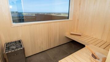 A sauna room with a window, a wooden bench, a small trash bin, and a wooden bucket.