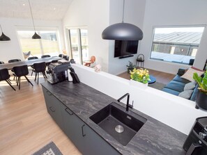 A modern kitchen with a large island, a dining area with chairs, a sofa, and a TV mounted on the wall. - 5 Star Holiday Home in Lemvig (Lemvig)