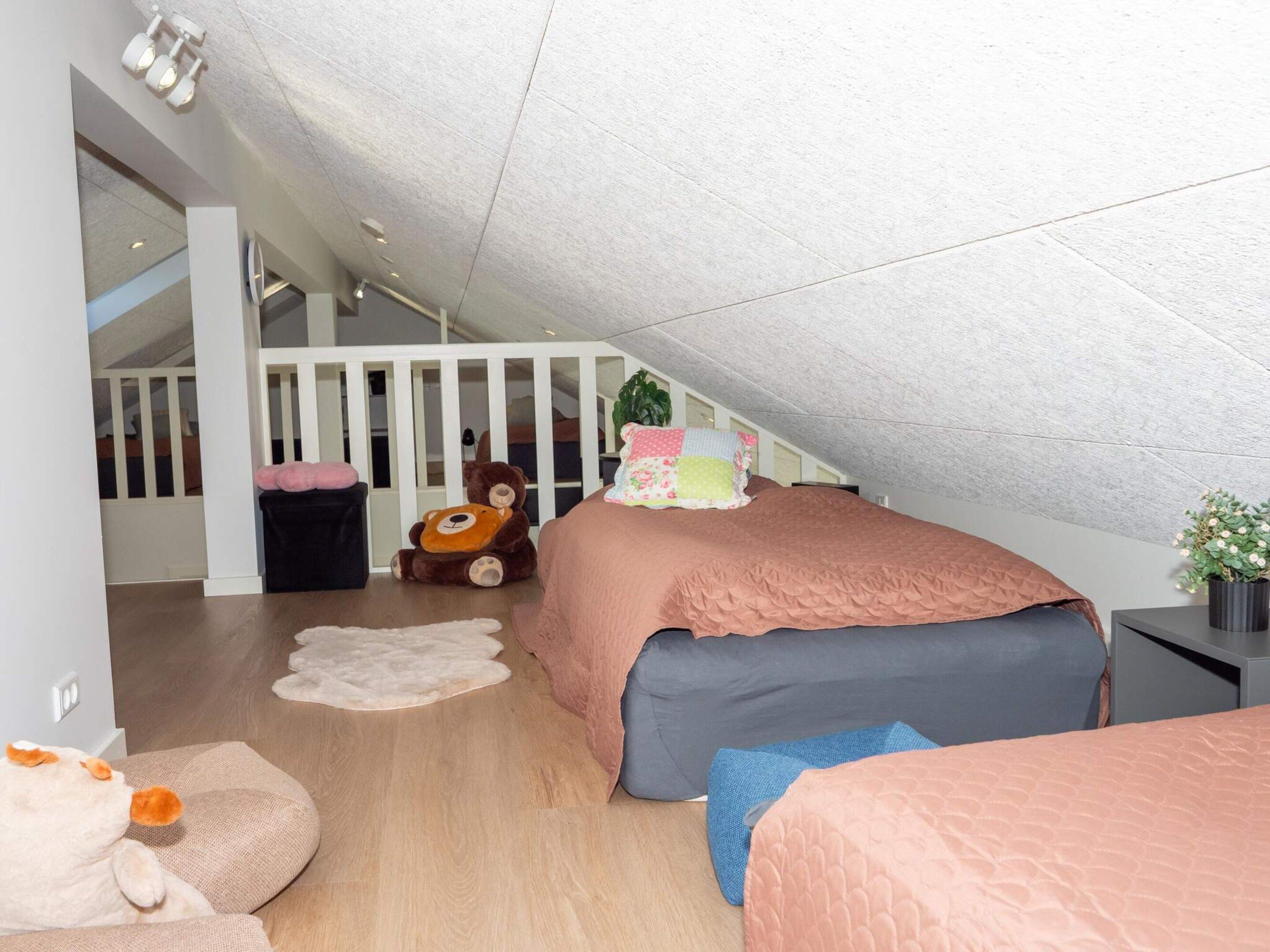A child's bedroom with a sloped ceiling, a bed with a pink and blue bedspread, a teddy bear, and a small potted plant.