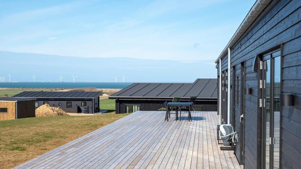 A modern building with a wooden deck, a table with chairs, and a view of wind turbines in the distance. - 5 Star Holiday Home in Lemvig (Lemvig)