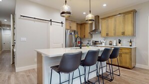 A modern kitchen with a central island, bar stools, wooden cabinets, stainless steel appliances, and pendant lighting. - River City Resorts: Rio Guad 313: Remarkable Condo, Guadalupe River View! (New Braunfels)