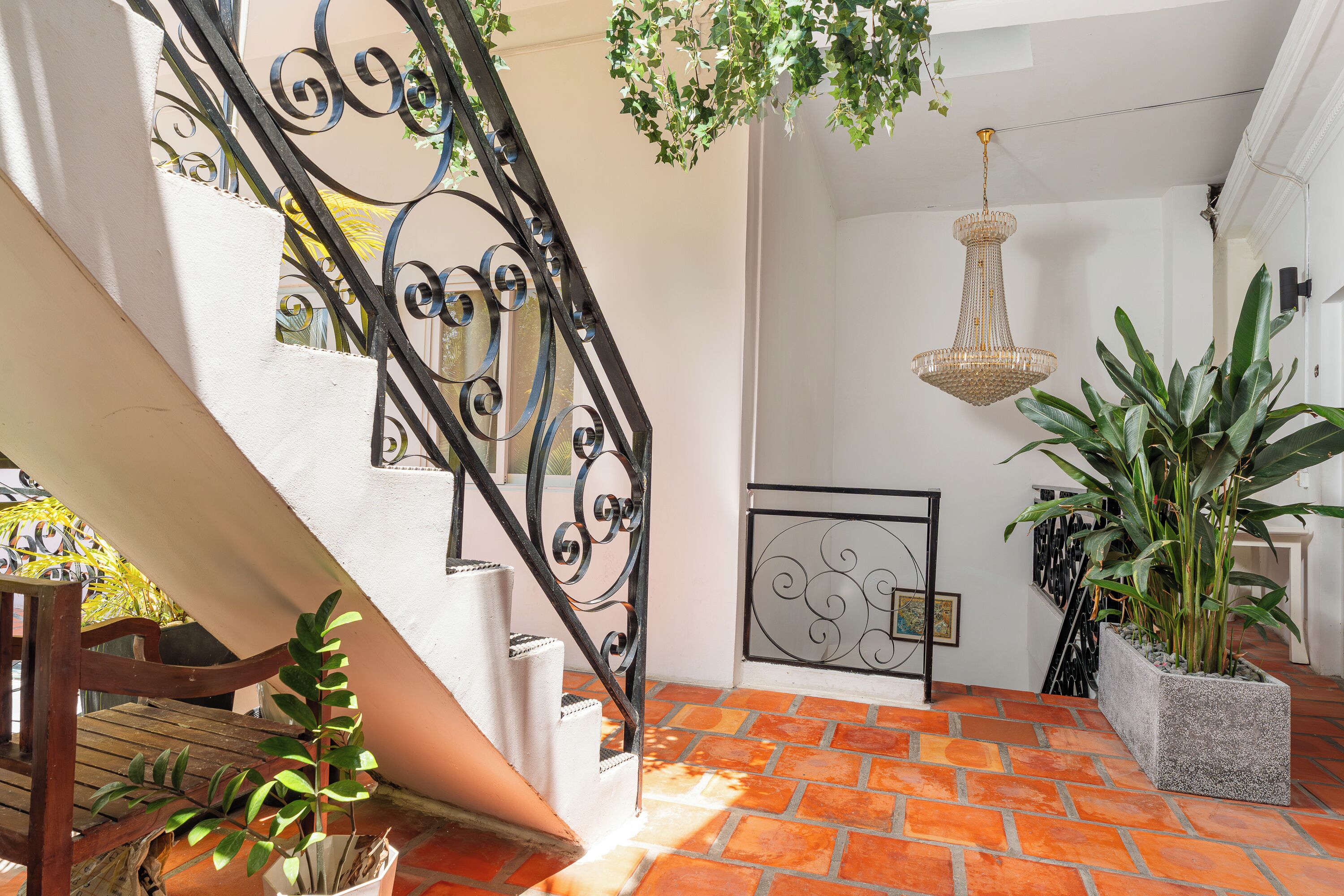Una escalera con barandales de metal negro ornamentados, un candelabro colgante y plantas en macetas.