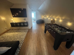 Tunnelmallinen huone, jossa on pöytäjalkapallopeli, seinälle asennettu televisio ja valoketju.